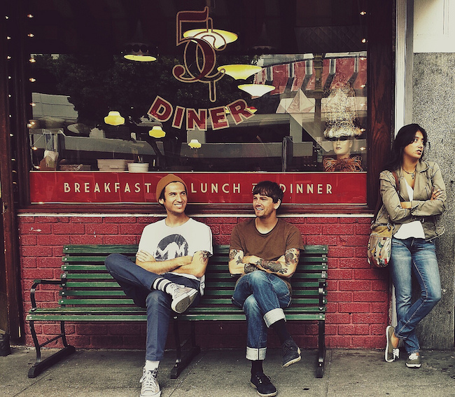 two men sitting on a bench outside a diner, a woman leans against a wall next to them